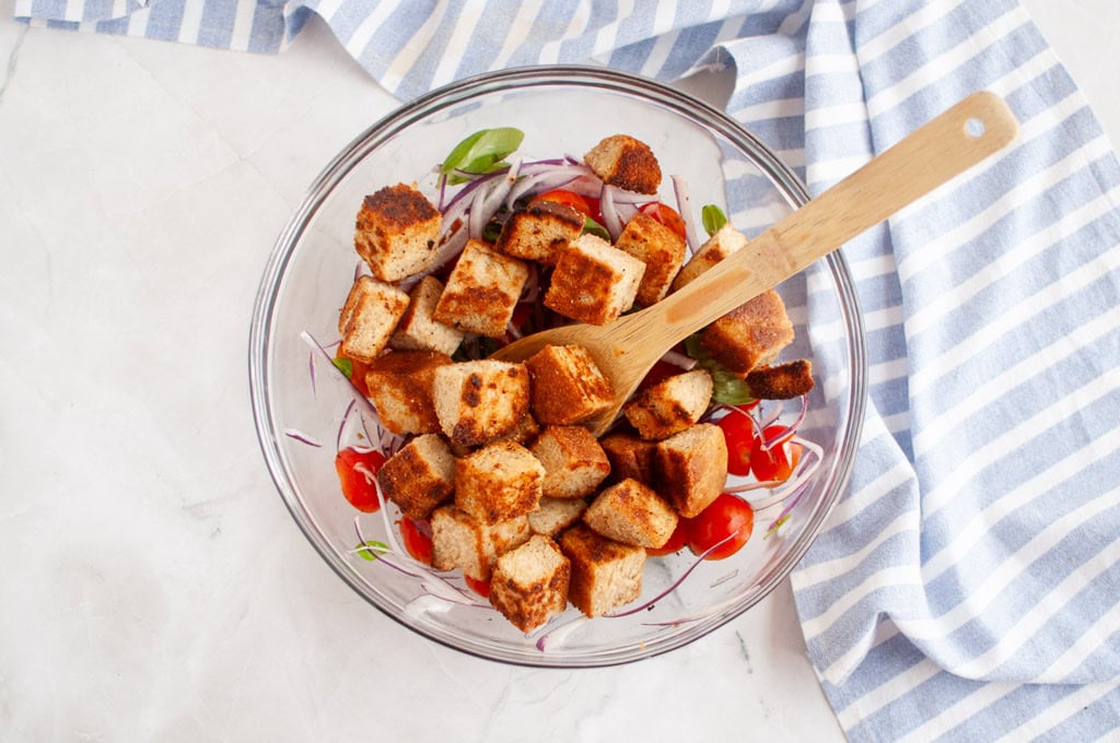 Overhead shot of gluten-free croutons, tomatoes, and red onion in a glass bowl, with a wooden spoon and striped cloth.