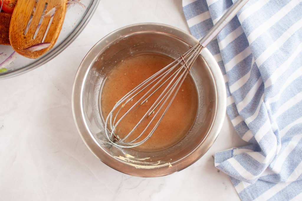 Overhead shot of a light-colored dressing in a metal bowl with a silver whisk, alongside a striped cloth and blurred ingredients.