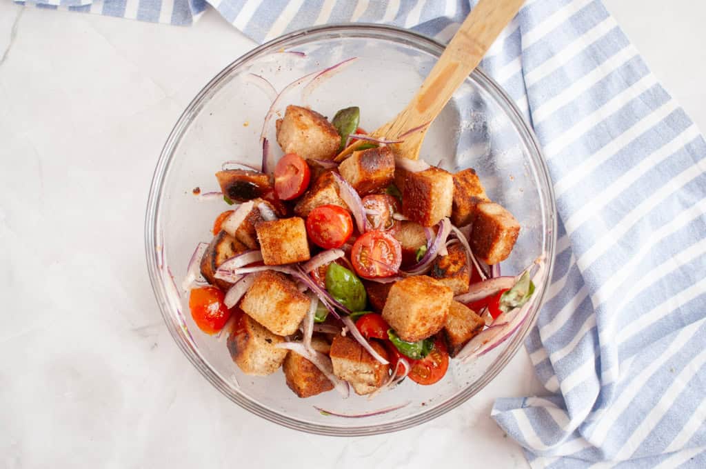 Overhead shot of panzanella salad in a glass bowl, featuring toasted bread cubes, cherry tomatoes, red onion, and basil leaves, with a wooden spoon.