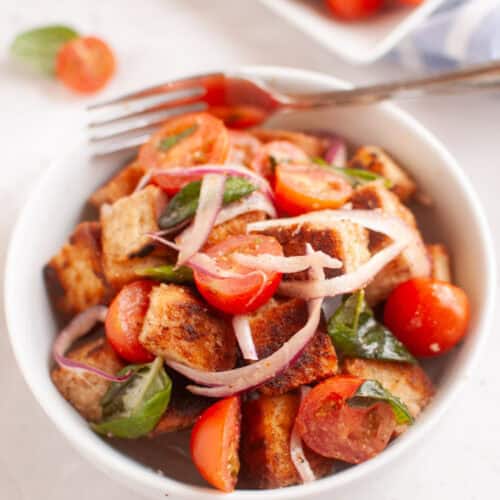 White bowl of panzanella salad with toasted bread cubes, cherry tomatoes, red onion, and basil leaves.