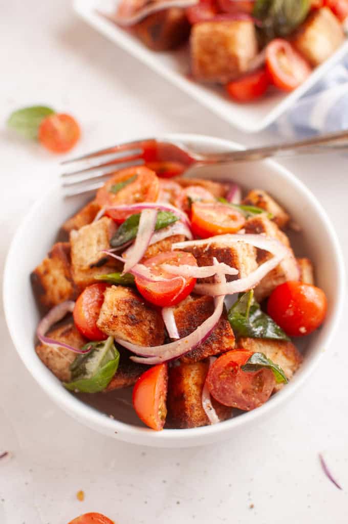 A white bowl filled with panzanella salad, featuring toasted bread cubes, cherry tomatoes, red onion, and basil leaves.