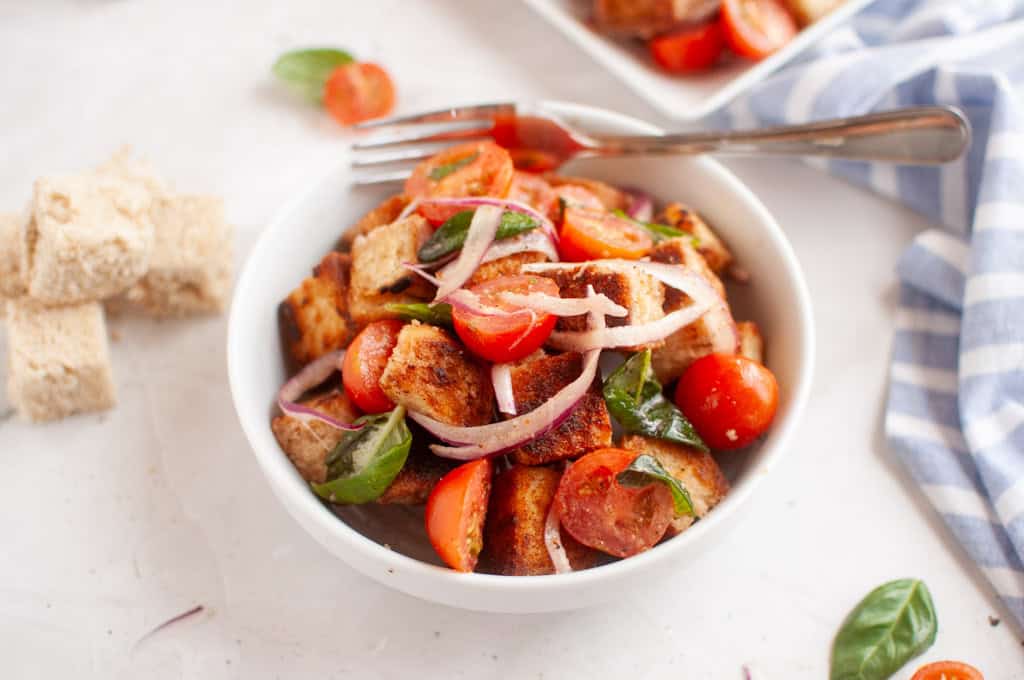 A white bowl filled with panzanella salad, featuring toasted bread cubes, cherry tomatoes, red onion, and basil leaves, is shown with a fork.