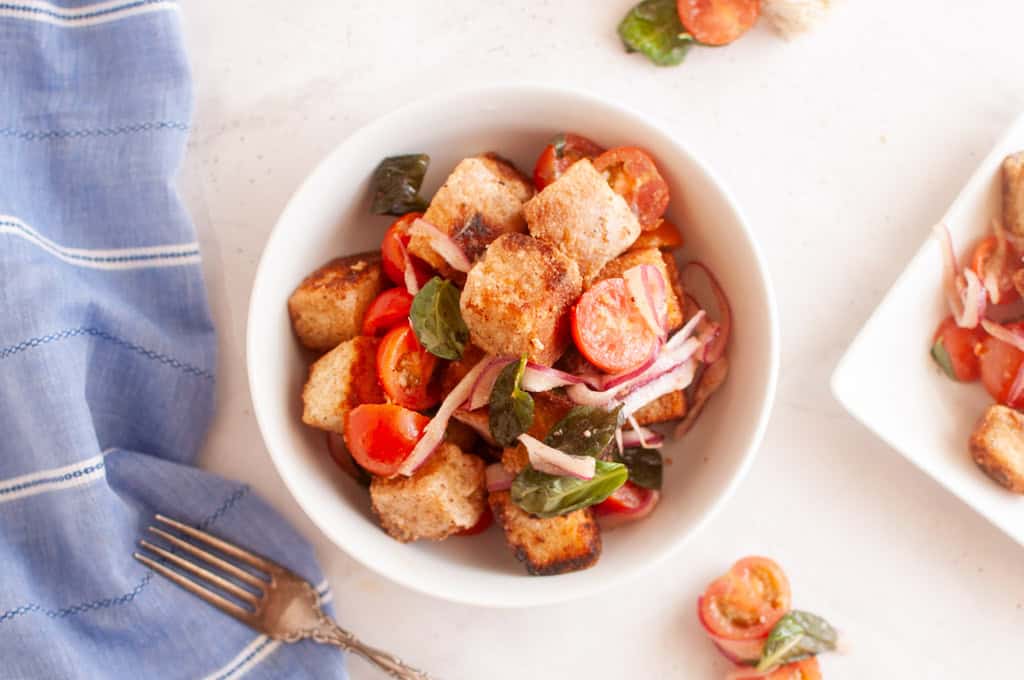 Overhead shot of panzanella salad in a white bowl, featuring toasted bread cubes, cherry tomatoes, red onion, and basil leaves, with a fork and cloth.