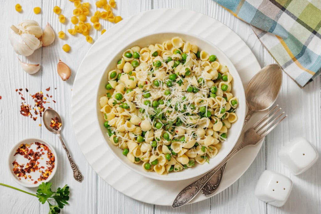 A white bowl filled with pasta and peas, garnished with cheese and parsley, is shown with spoons, garlic, and spices.