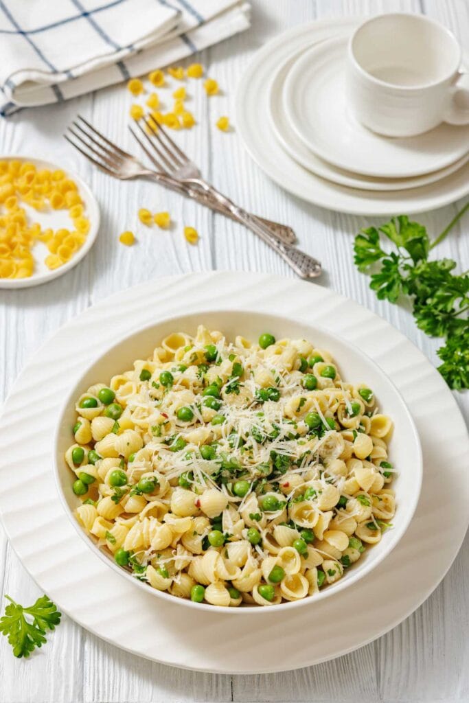A white bowl filled with pasta and peas, garnished with cheese and parsley, is shown with forks and plates.