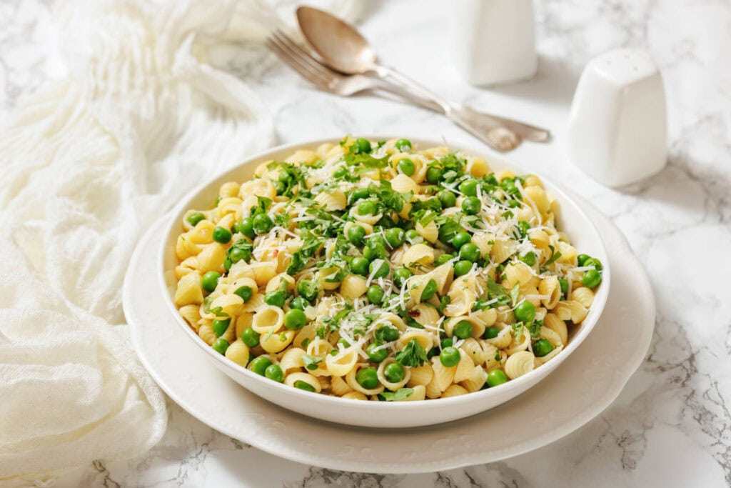 A white bowl filled with pasta and peas, garnished with cheese and parsley, is shown with spoons and salt and pepper shakers.