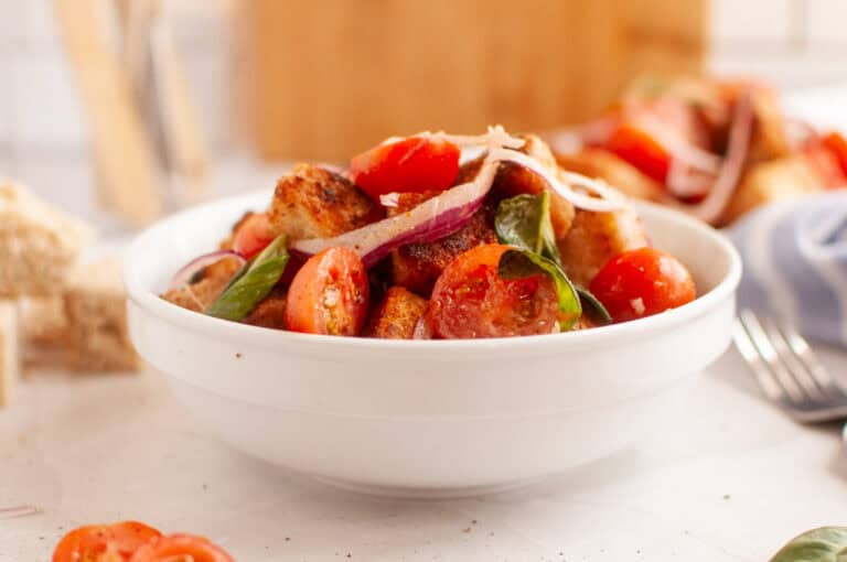 A white bowl filled with panzanella salad, featuring toasted bread cubes, cherry tomatoes, red onion, and basil leaves.