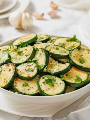 A white bowl filled with sliced, cooked zucchini garnished with chopped herbs, placed on a white table with a fork.