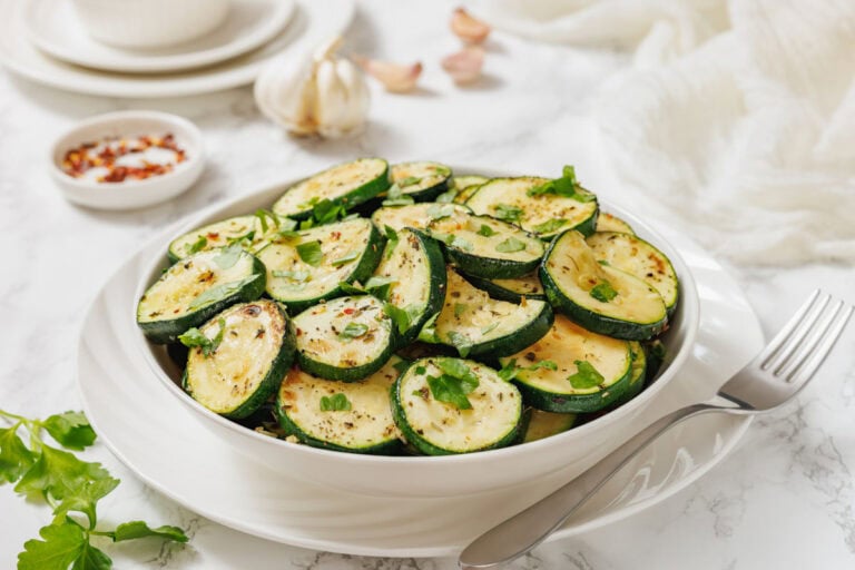 A white bowl filled with sliced, cooked zucchini garnished with chopped herbs, placed on a white table with a fork.