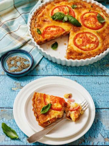 A slice of tomato pie on a white plate with a fork, and a whole tomato pie in a white fluted dish, are shown on a blue wooden surface with a striped cloth and a small bowl of spices.