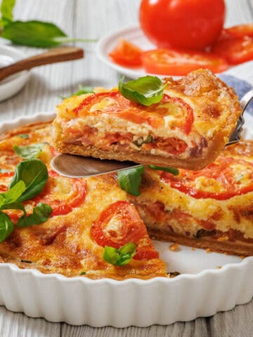 A slice of tomato pie being lifted with a serving utensil from a white fluted dish. A whole tomato pie is visible in the dish, garnished with tomato slices and basil.