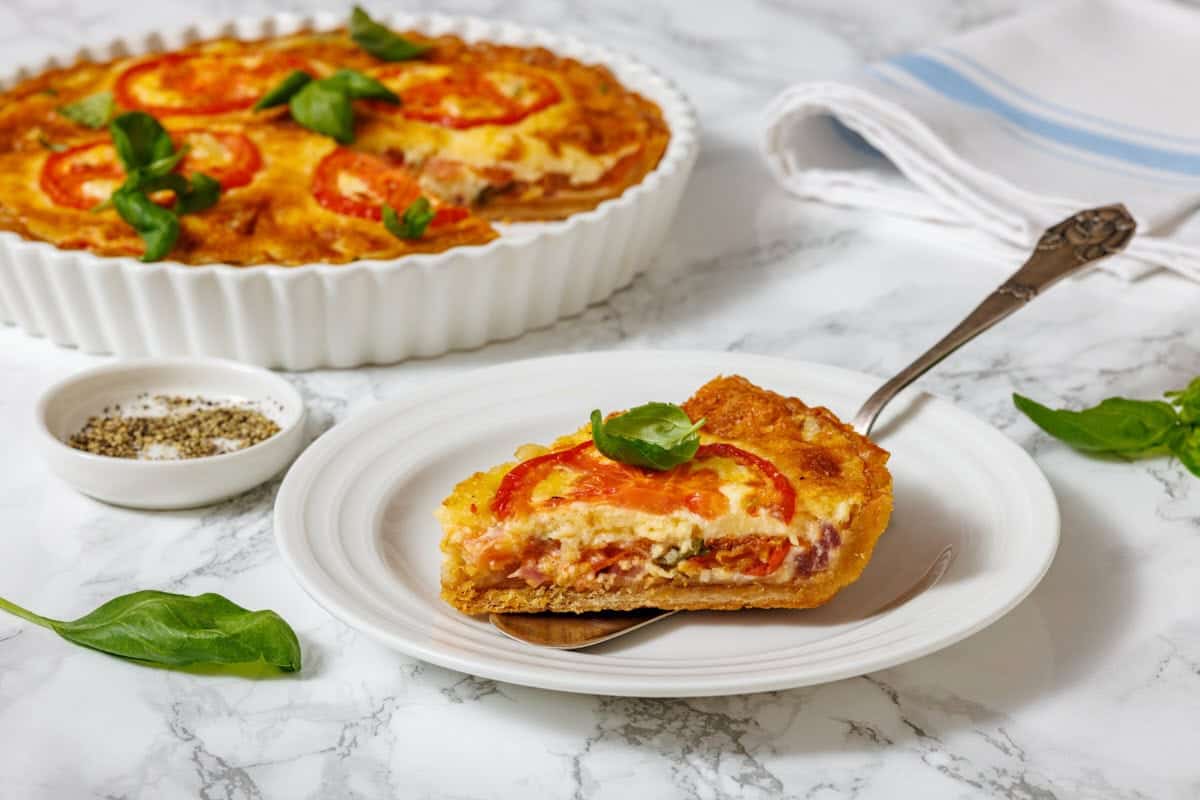 A slice of tomato pie on a white plate with a serving utensil, and a whole tomato pie in a white fluted dish, are shown on a marble surface with a small bowl of spices and basil leaves.