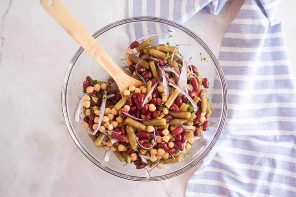 A glass bowl filled with a Three Bean Salad including kidney beans, chickpeas, green beans, and sliced red onion, with a wooden spoon and a striped cloth nearby.