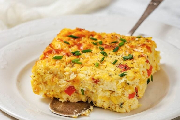 A square slice of Corn Pudding Casserole with red peppers and chopped chives sits on a white plate with a serving spatula.
