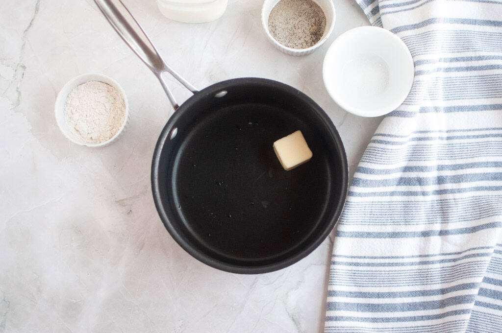 Overhead shot of a pat of butter melting in a black saucepan, surrounded by ingredients for creamed peas.