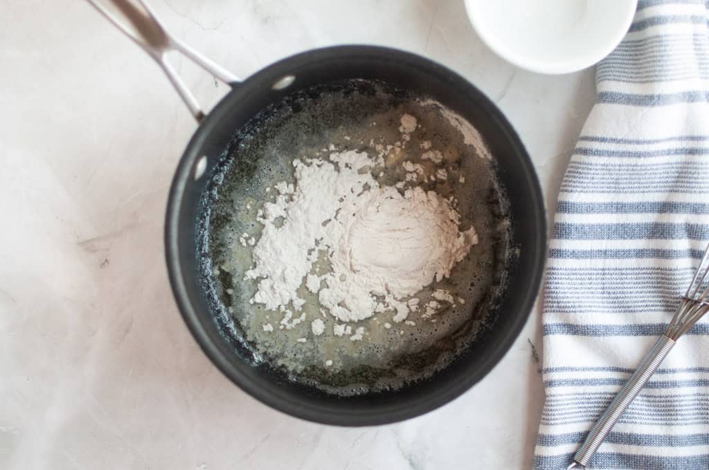 Overhead shot of flour being added to melted butter in a black saucepan, with other ingredients nearby.