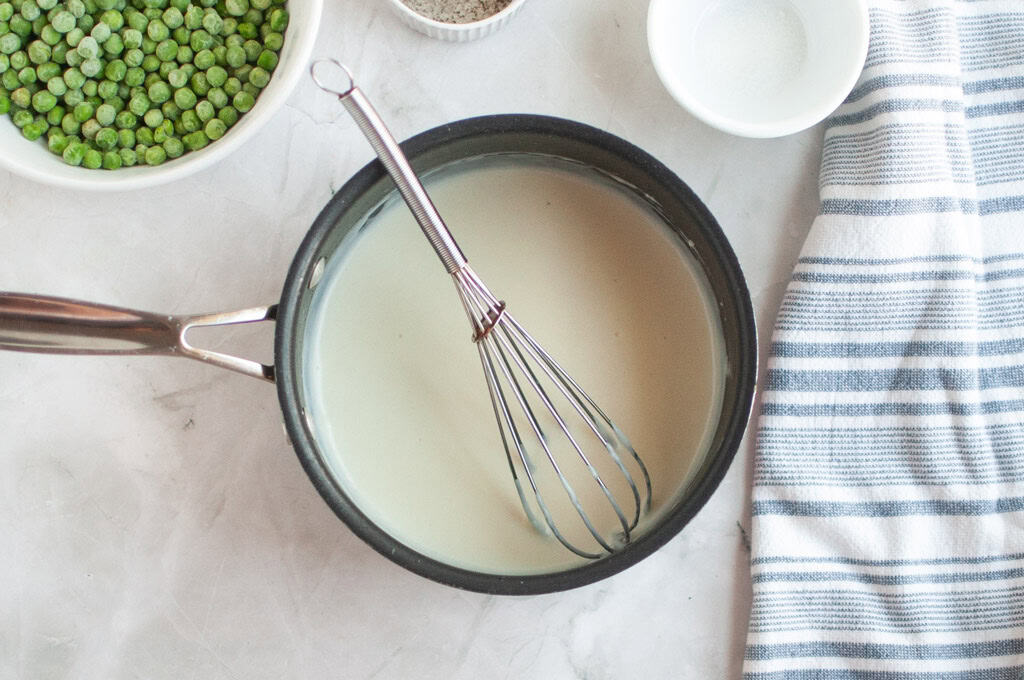 Overhead shot of a creamy white B&eacute;chamel sauce being whisked in a black saucepan, with peas and pepper nearby.