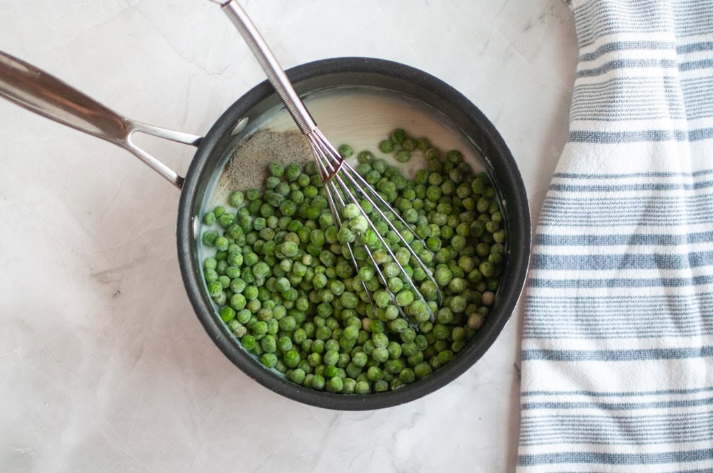 Overhead shot of frozen green peas, milk, and black pepper in a black saucepan being stirred with a whisk.