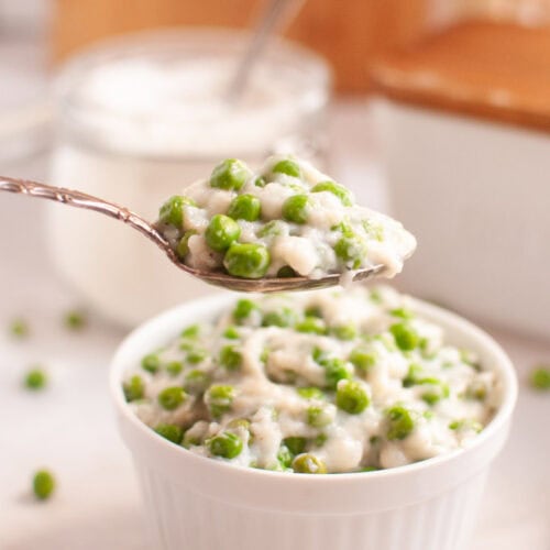 A spoonful of creamy green peas is held above a white bowl filled with the same dish.