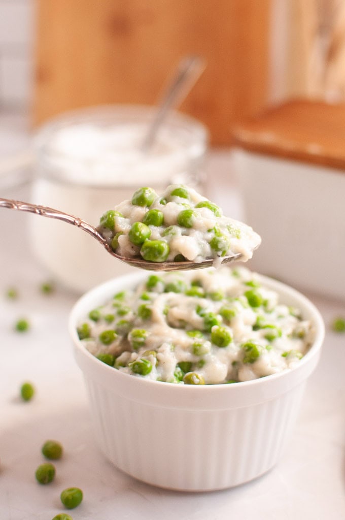 A spoonful of creamy green peas is held above a white bowl filled with the same dish.