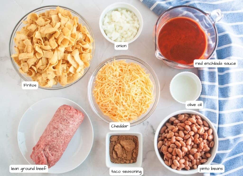 Overhead shot of ingredients for Frito pie, including Fritos, onions, enchilada sauce, cheese, ground beef, spices, oil, and beans, arranged on a light surface with a blue striped cloth and labels for each ingredient.