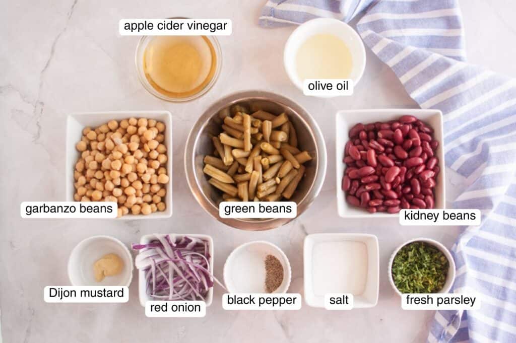 Overhead view of labeled ingredients for Grandma&rsquo;s Three Bean Salad, including garbanzo beans, green beans, kidney beans, red onion, Dijon mustard, black pepper, salt, fresh parsley, apple cider vinegar, and olive oil arranged on a light surface with a striped cloth.