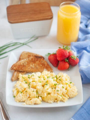A plate with scrambled eggs, toast, and strawberries next to a glass of orange juice on a table with a blue napkin.