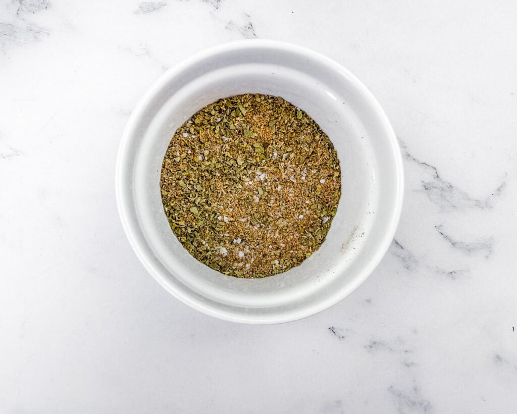 Overhead shot of a white bowl containing a mixture of dried herbs and spices, resting on a white marble surface with gray veins.