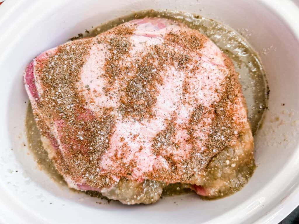 Overhead shot of a raw pork roast seasoned with spices, sitting in a white slow cooker with a thin liquid at the bottom. The pork has a pinkish hue and is coated with a visible blend of spices.