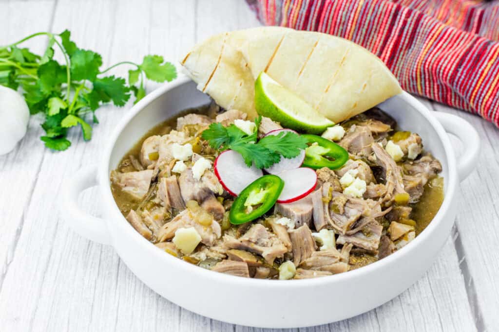 A white bowl of Slow Cooker Pork Chile Verde, featuring visible chunks of meat, radish slices, jalapeno slices, cilantro, and cheese, sits on a white wooden table. A folded tortilla and lime wedge are placed beside the bowl. A head of garlic and cilantro sprigs are in the background, along with a red and white striped cloth.
