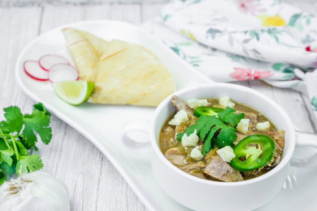 A white bowl of Slow Cooker Pork Chile Verde, featuring visible chunks of meat, white cheese crumbles, sliced jalapenos, and cilantro, sits on a white plate. Sliced radishes, a lime wedge, and folded tortillas are also on the plate. A head of garlic and cilantro sprigs are in the blurred background, along with a floral patterned cloth.