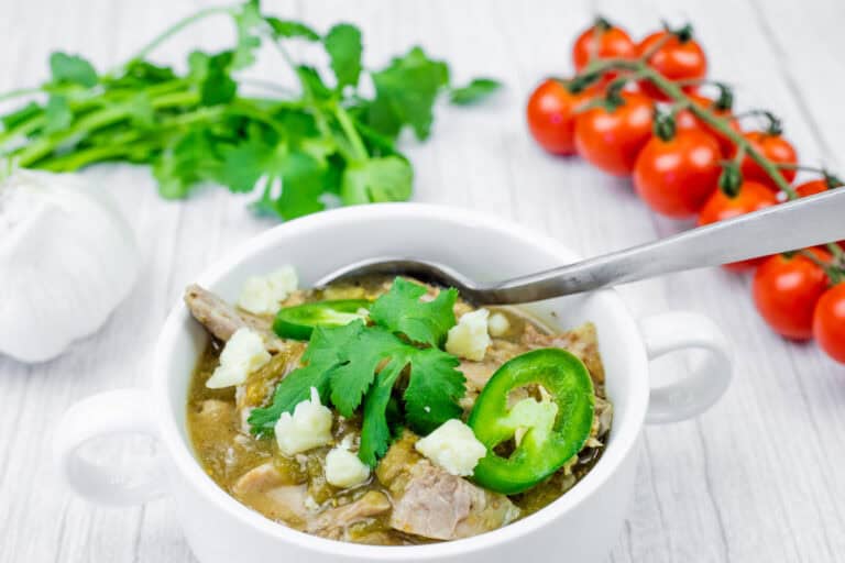 Bowl of slow cooker pork chile verde with toppings and a spoon on a white wooden table.
