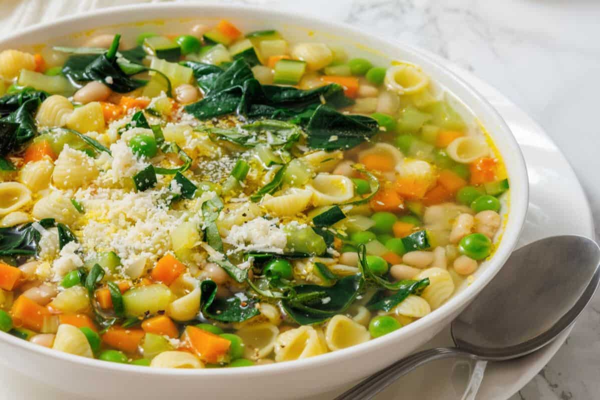 A bowl of Spring Minestrone sits on a plate with a spoon beside it.