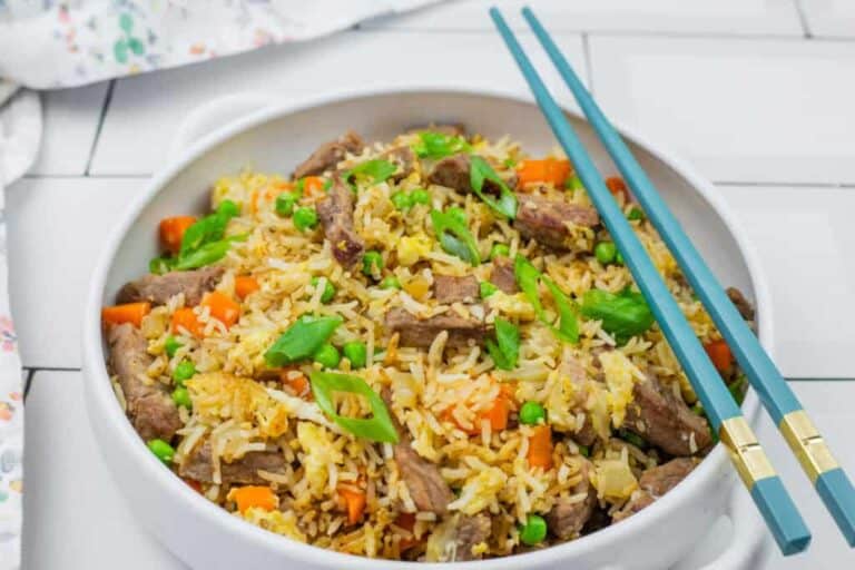 A bowl of steak fried rice served with blue chopsticks on a white tiled surface.
