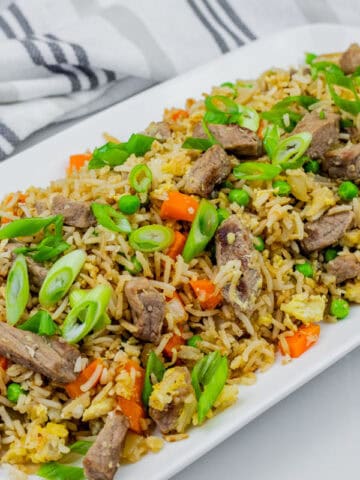 Steak fried rice on a white platter on white tiles with striped cloth on the side.