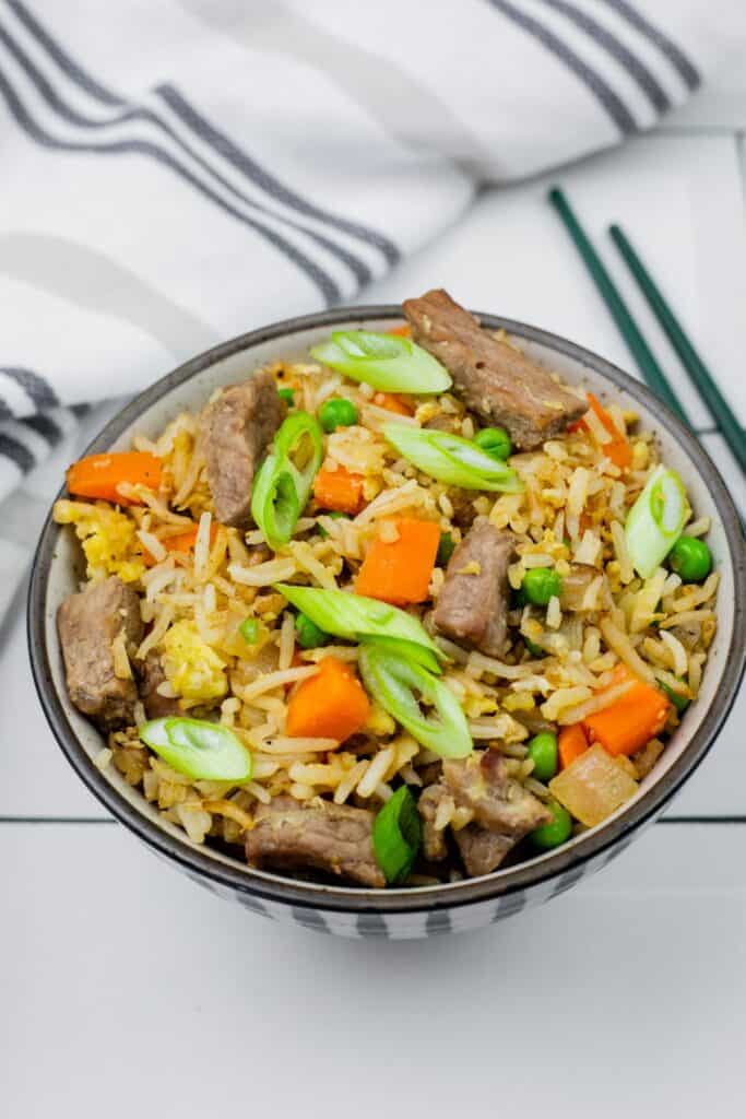 Steak fried rice with green onions and carrots in a bowl and a striped cloth nearby.