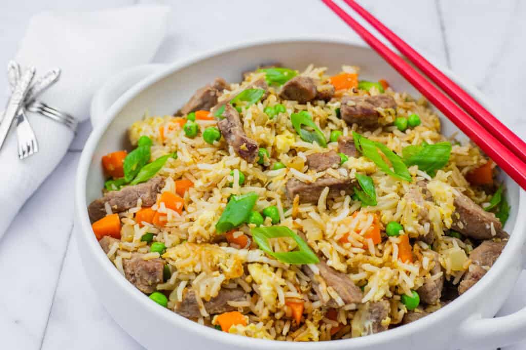 steak fried rice in a white bowl with handles and red-colored chopsticks on top.