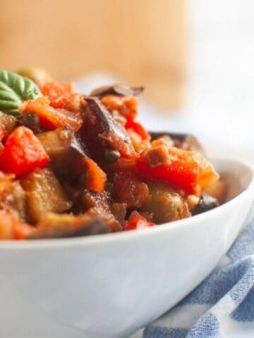 A white bowl filled with a vegetable stew containing tomatoes, eggplant, and other vegetables, garnished with a basil leaf.