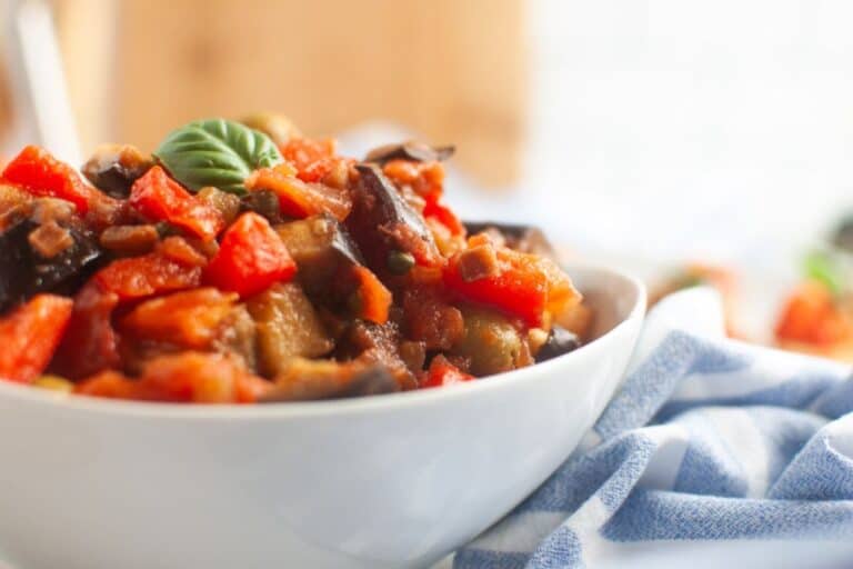 A white bowl filled with a vegetable stew containing tomatoes, eggplant, and other vegetables, garnished with a basil leaf.