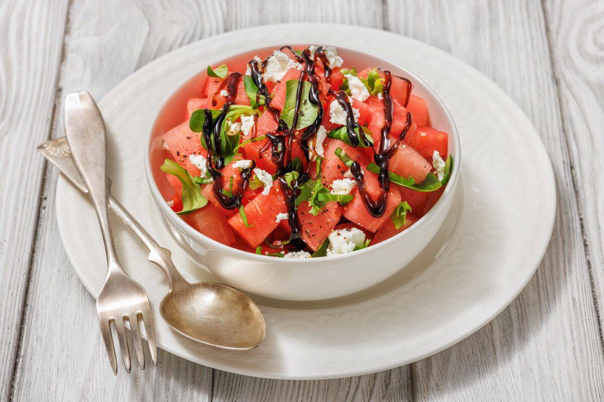 A bowl of watermelon salad with feta cheese, arugula, and balsamic glaze, placed on a white plate with a fork and spoon on the side.