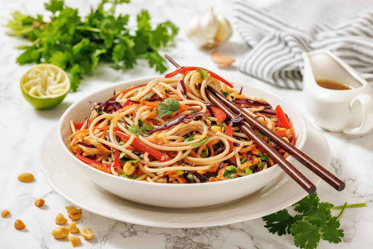 A bowl of Asian-inspired noodle salad with vegetables, garnished with cilantro, chopsticks on top, and ingredients like lime, peanuts, and fresh herbs nearby.