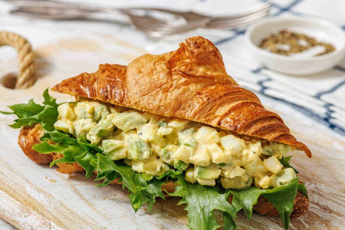 A croissant sandwich filled with Avocado egg salad, lettuce, and herbs sits on a wooden board.