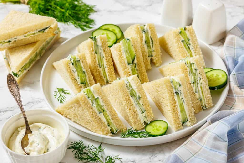 A plate of neatly arranged cucumber tea sandwiches on white bread, garnished with fresh dill and cucumber slices, with a bowl of creamy dip on the side.