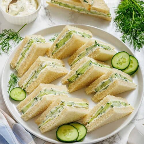 A plate of neatly arranged cucumber tea sandwiches with cream cheese, garnished with fresh dill and cucumber slices, sits on a marble surface beside a bowl of spread.