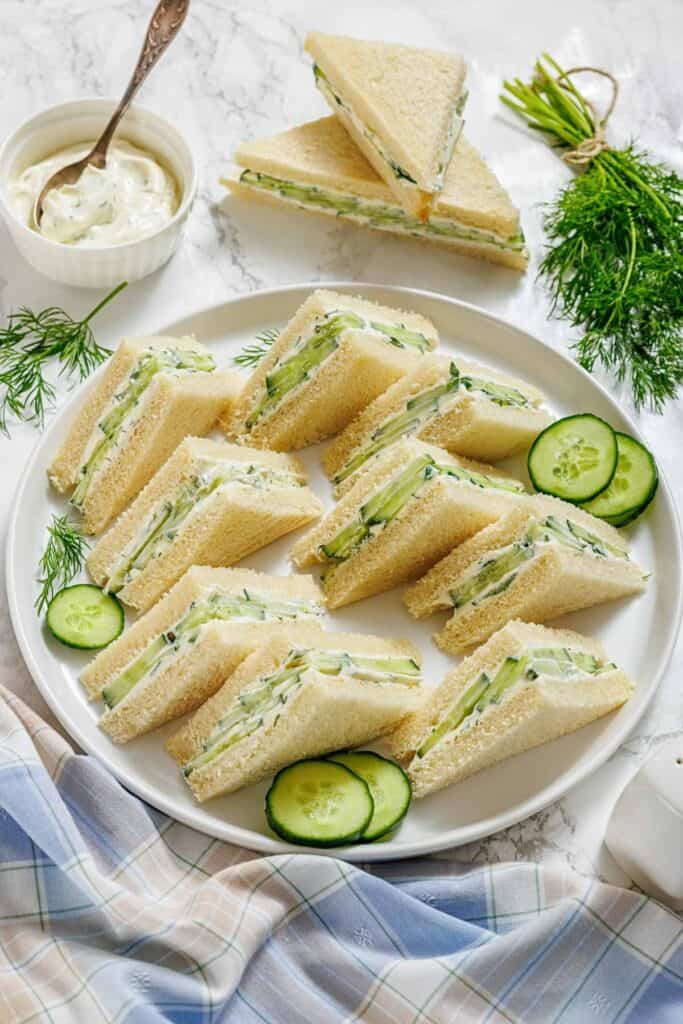 A plate of neatly arranged cucumber tea sandwiches with cream cheese, garnished with fresh dill and cucumber slices, sits on a marble surface beside a bowl of spread.