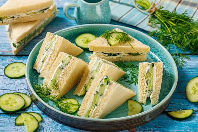A plate of triangular cucumber sandwiches on white bread, garnished with fresh dill and cucumber slices.