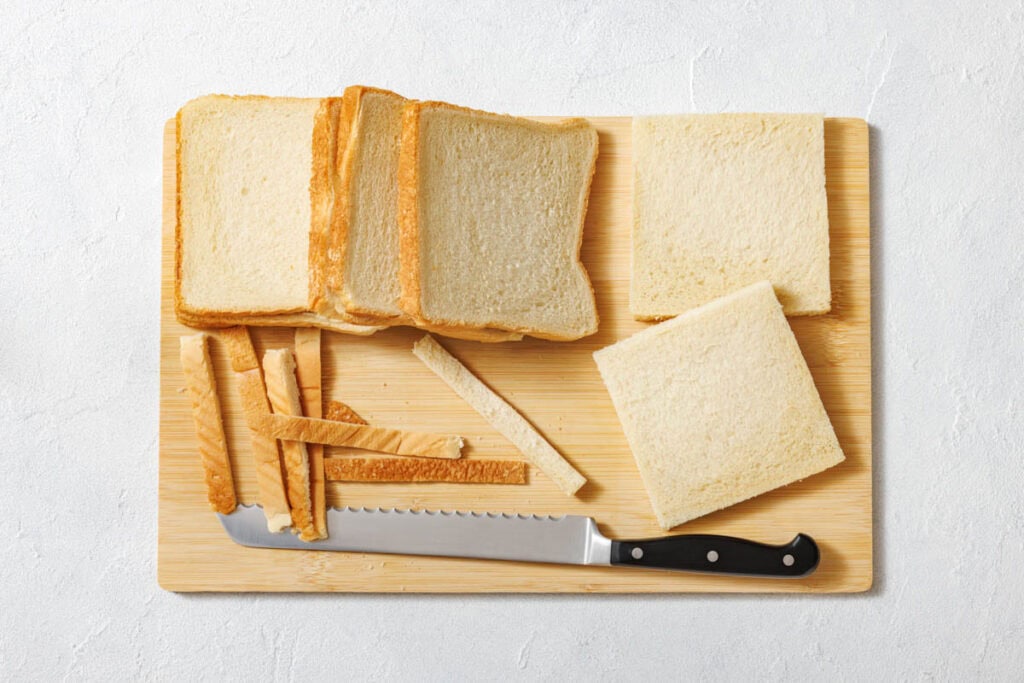 Slices of white bread and crusts on a wooden cutting board with a bread knife, viewed from above.