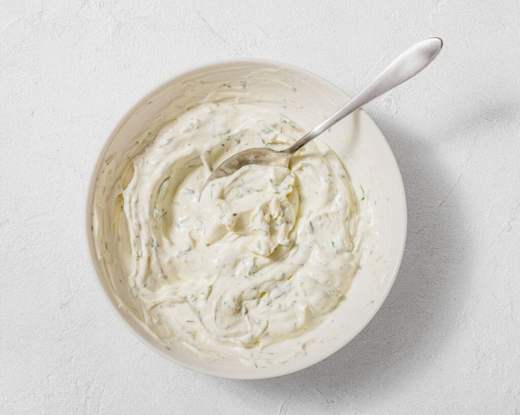A bowl of creamy white dip with green herb flecks, stirred with a metal spoon, on a light surface.
