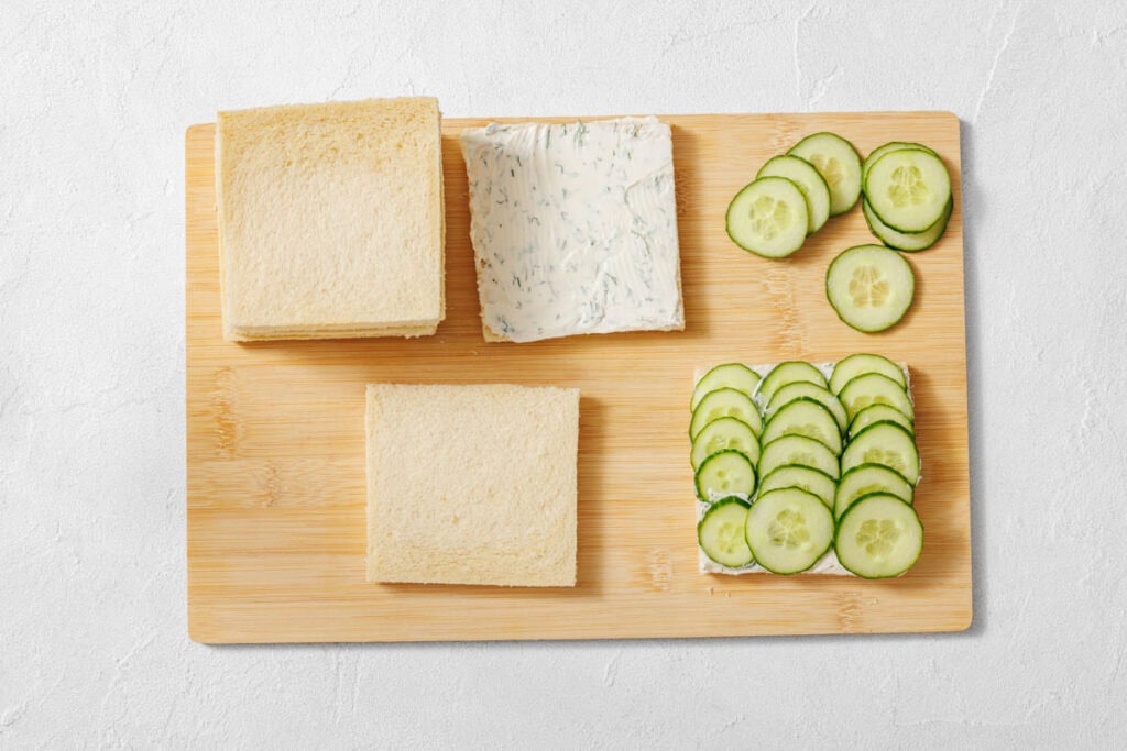 A wooden board with white bread slices, herbed cream cheese, and sliced cucumbers arranged for making sandwiches.