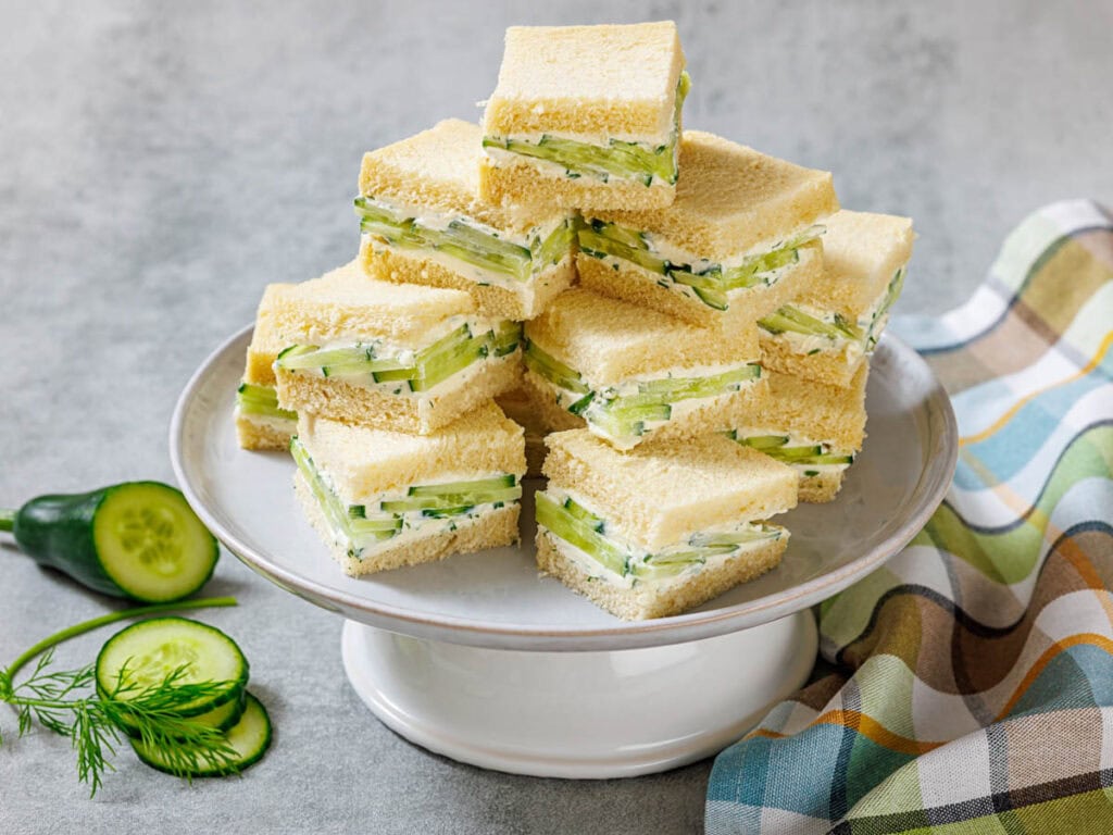 A plate of stacked cucumber tea sandwiches made with white bread, cream cheese, and sliced cucumbers, with herbs and a cloth napkin nearby.